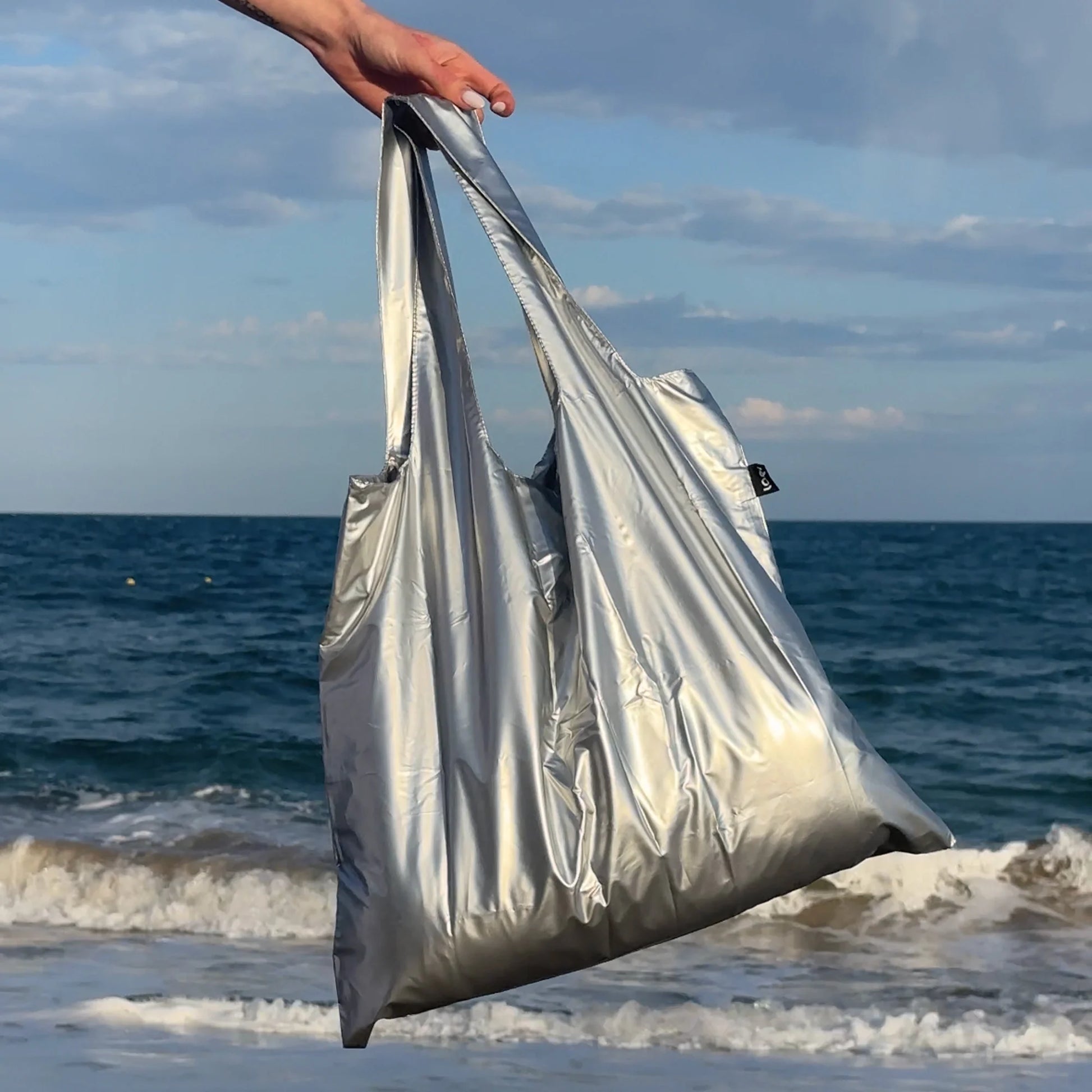 Torba LOQI metallic srebrna na plaży trzymana przez rękę z widokiem na morze i niebo