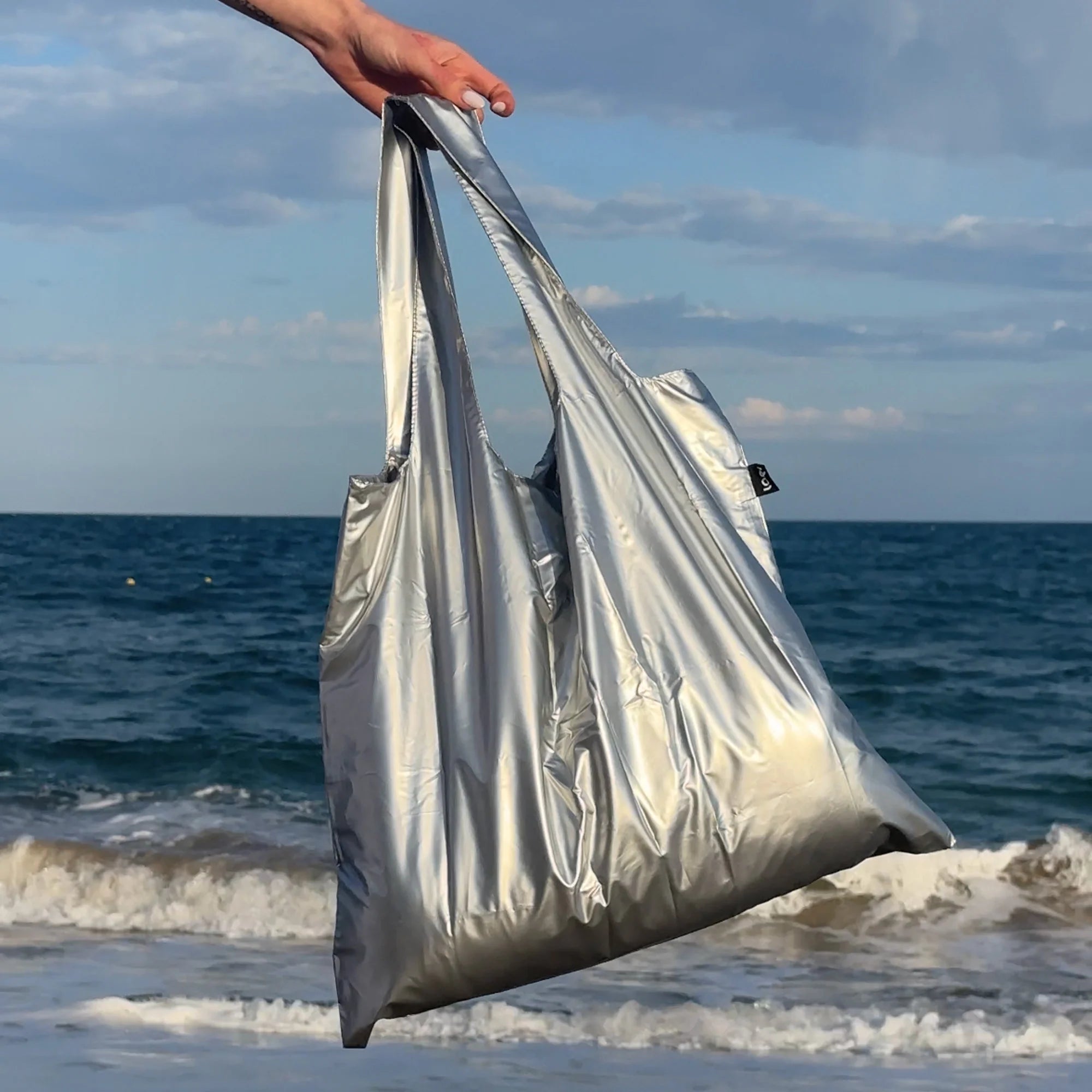 Torba LOQI metallic srebrna na plaży trzymana przez rękę z widokiem na morze i niebo