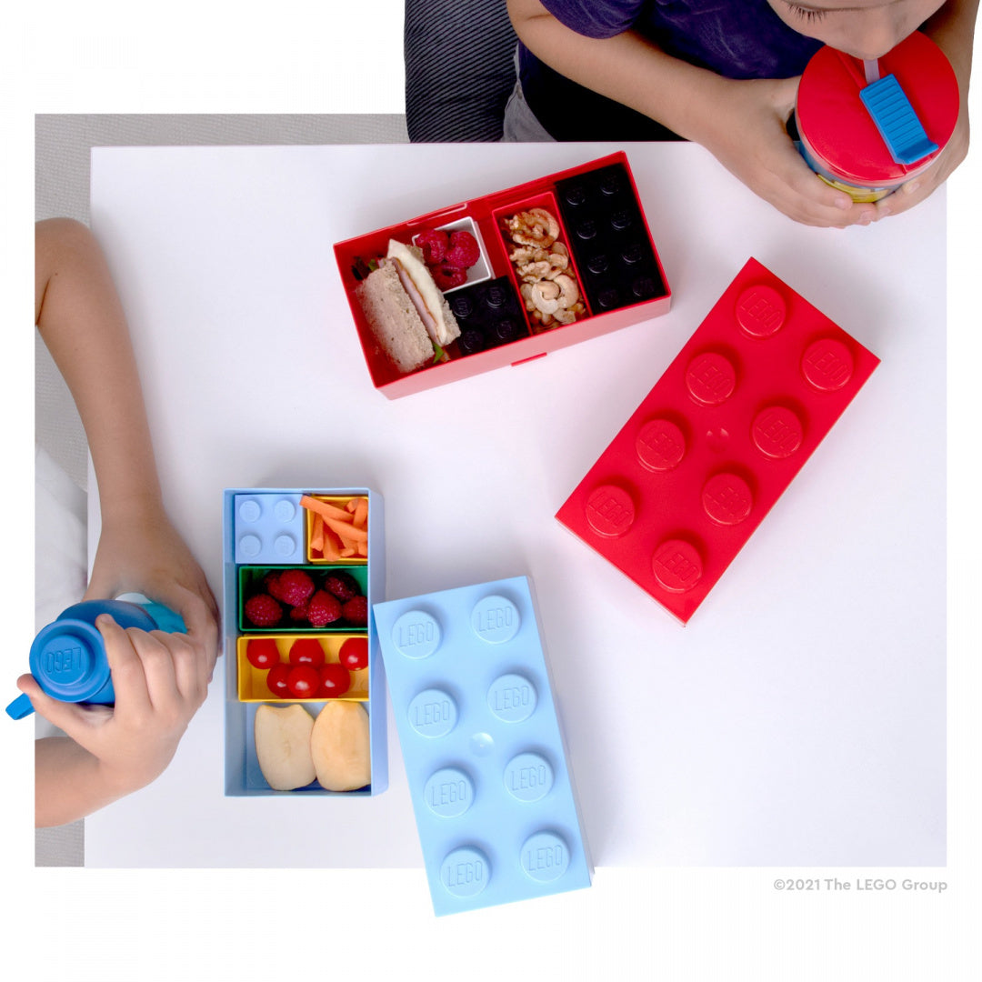 Lunchbox klocek LEGO jasnoniebieski z przegrodami na jedzenie obok czerwonego lunchboxa w kształcie klocka