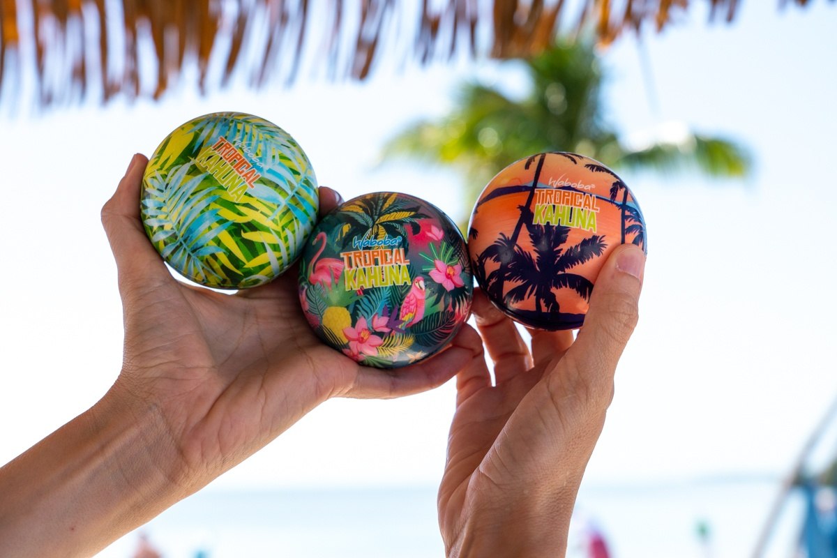 Trzy kolorowe piłki na wodę Waboba Tropical Kahuna z motywem tropikalnym trzymane w dłoniach na plaży