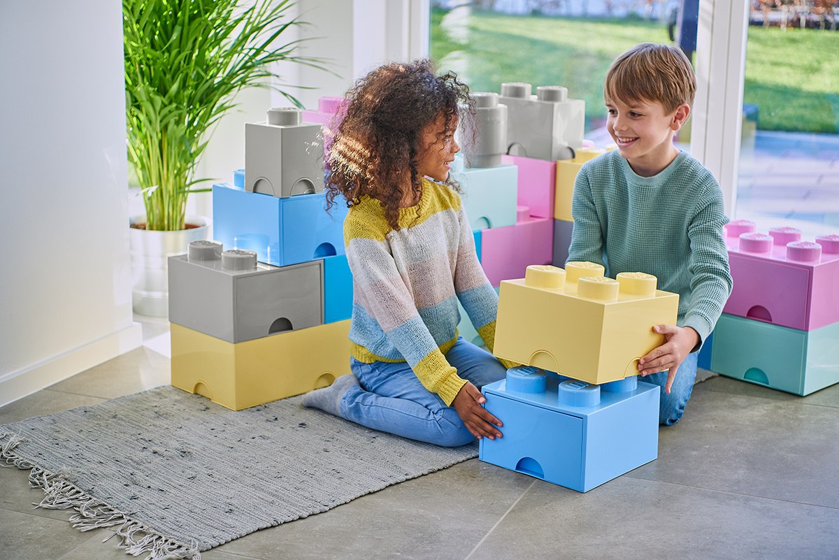 Dwoje dzieci bawiących się szarym pojemnikiem LEGO Brick szary oraz kolorowymi pojemnikami klocków w pokoju