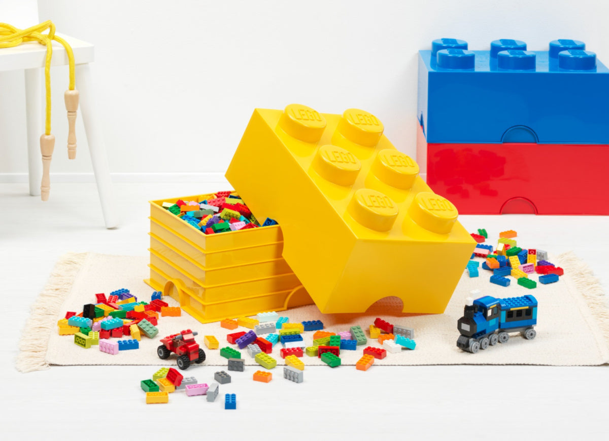 Żółty pojemnik LEGO na klocki w kształcie klocka z rozrzuconymi kolorowymi klockami na białym dywanie