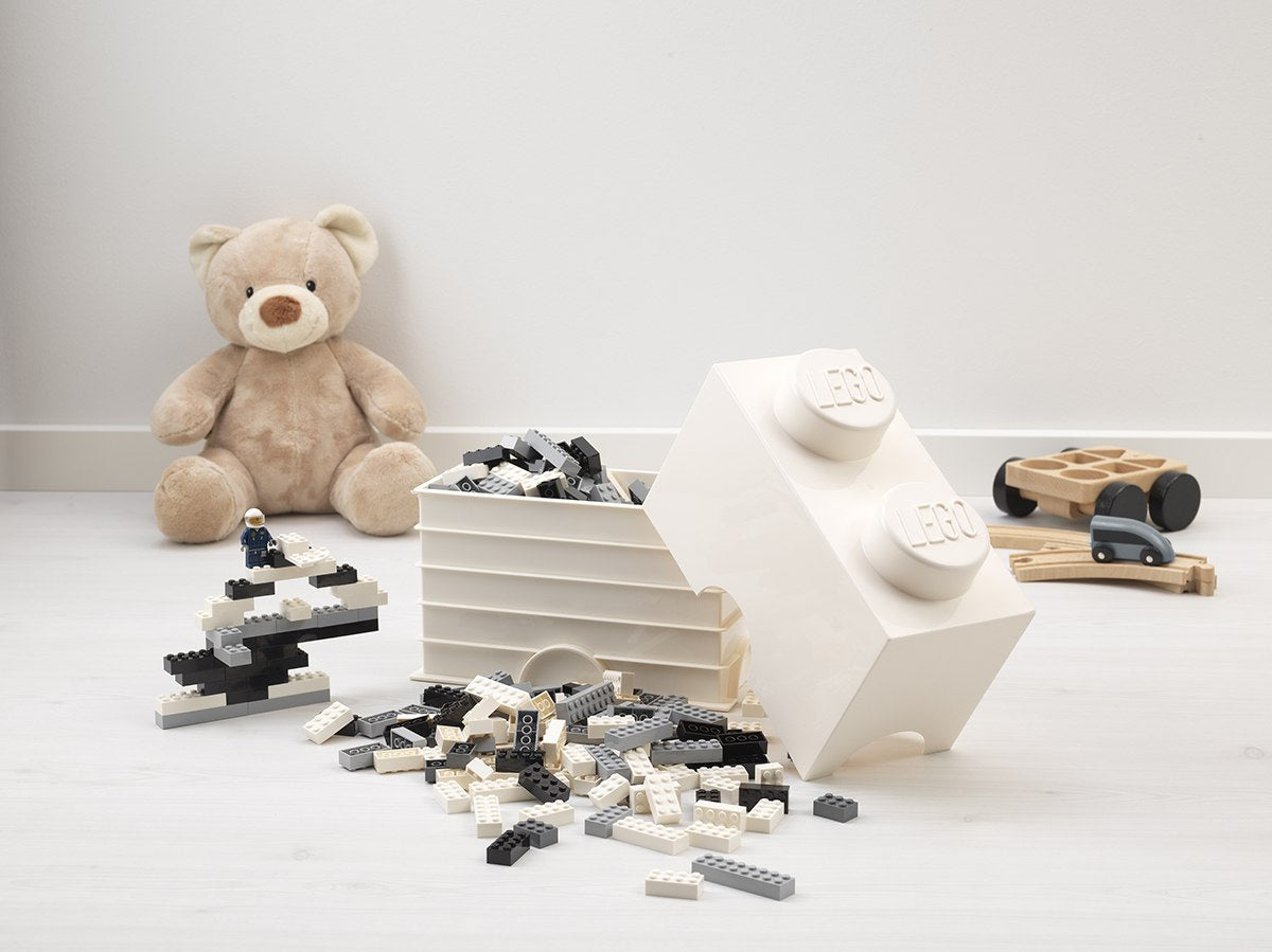 Pojemnik LEGO biały w kształcie dużej cegiełki z czarno-białymi klockami na podłodze i pluszowym misiem w tle
