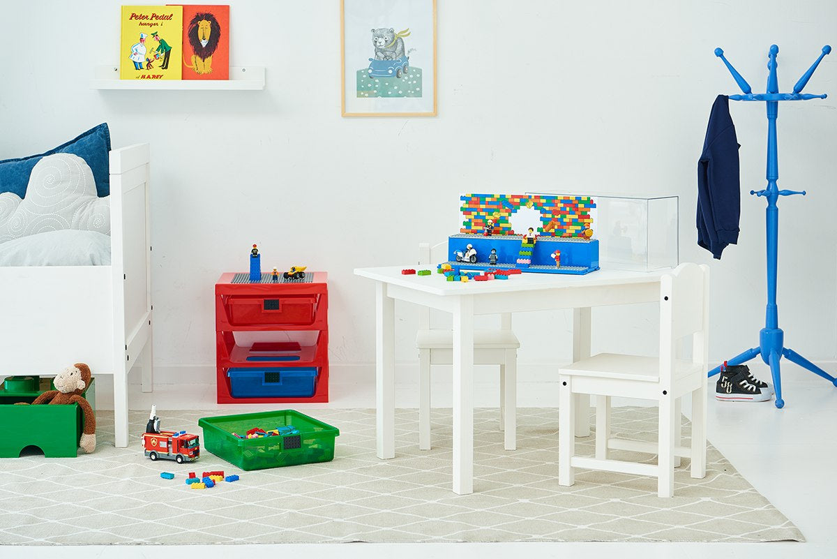 Biały pokój dziecięcy z regałem LEGO z szufladami w kolorach czerwonym i niebieskim obok białego stolika i krzesła