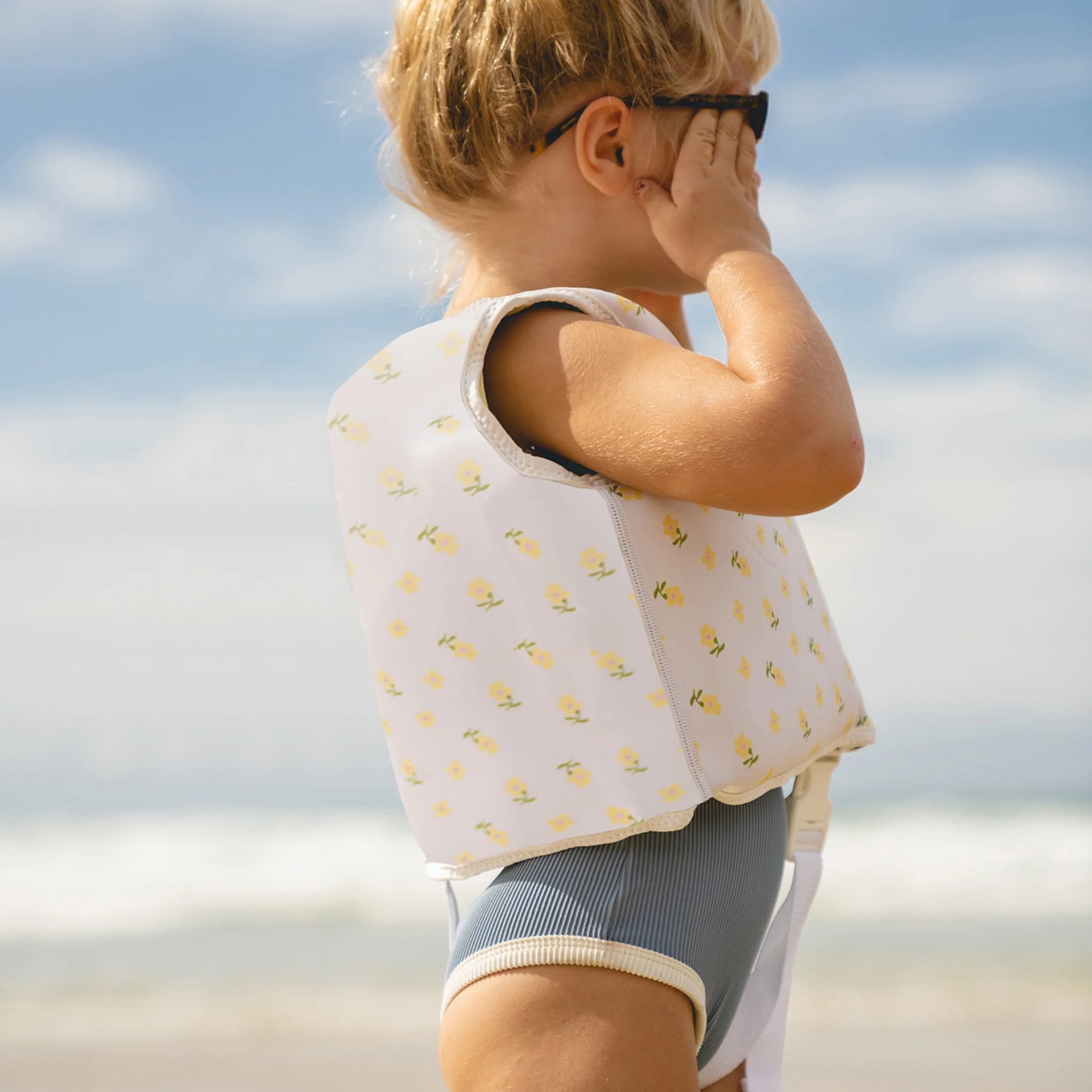 Dziecko na plaży w kwiatowej kamizelce do pływania dla dzieci marki SUNNY LIFE