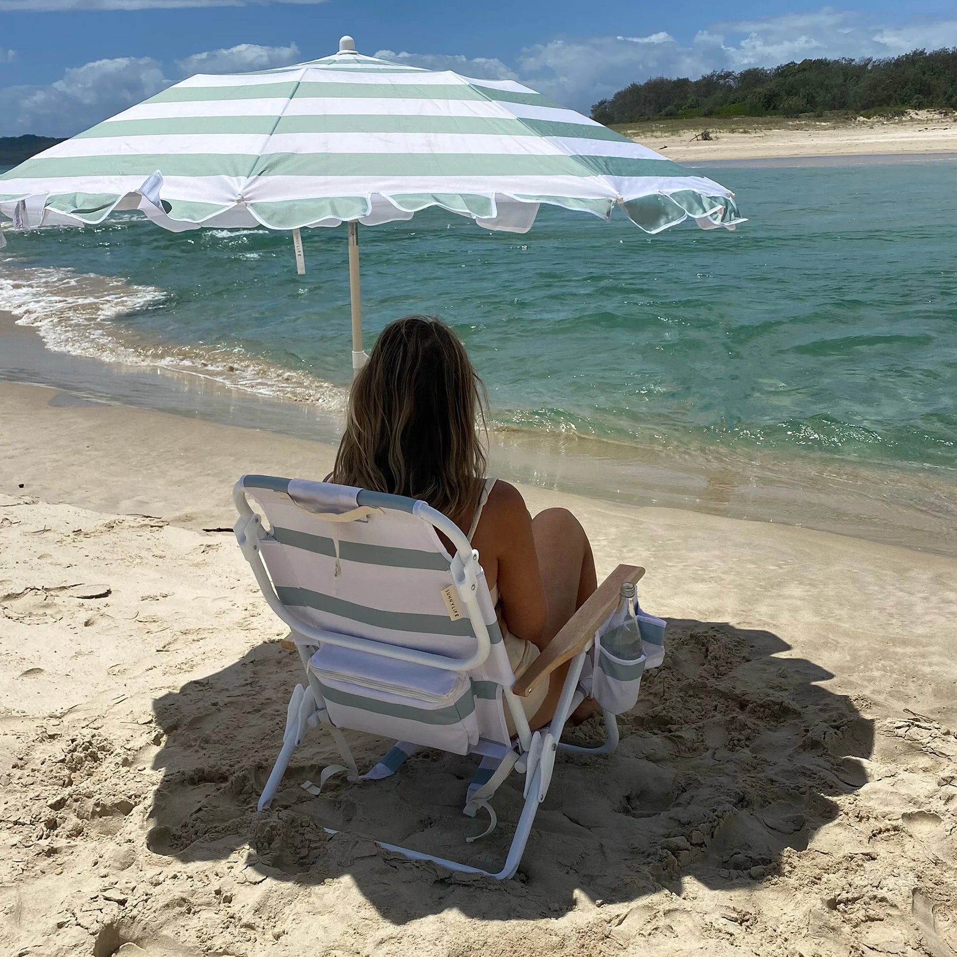 Kobieta siedząca na plaży pod zielono-białym kompaktowym parasolem plażowym LOQI z rozkładanym krzesłem