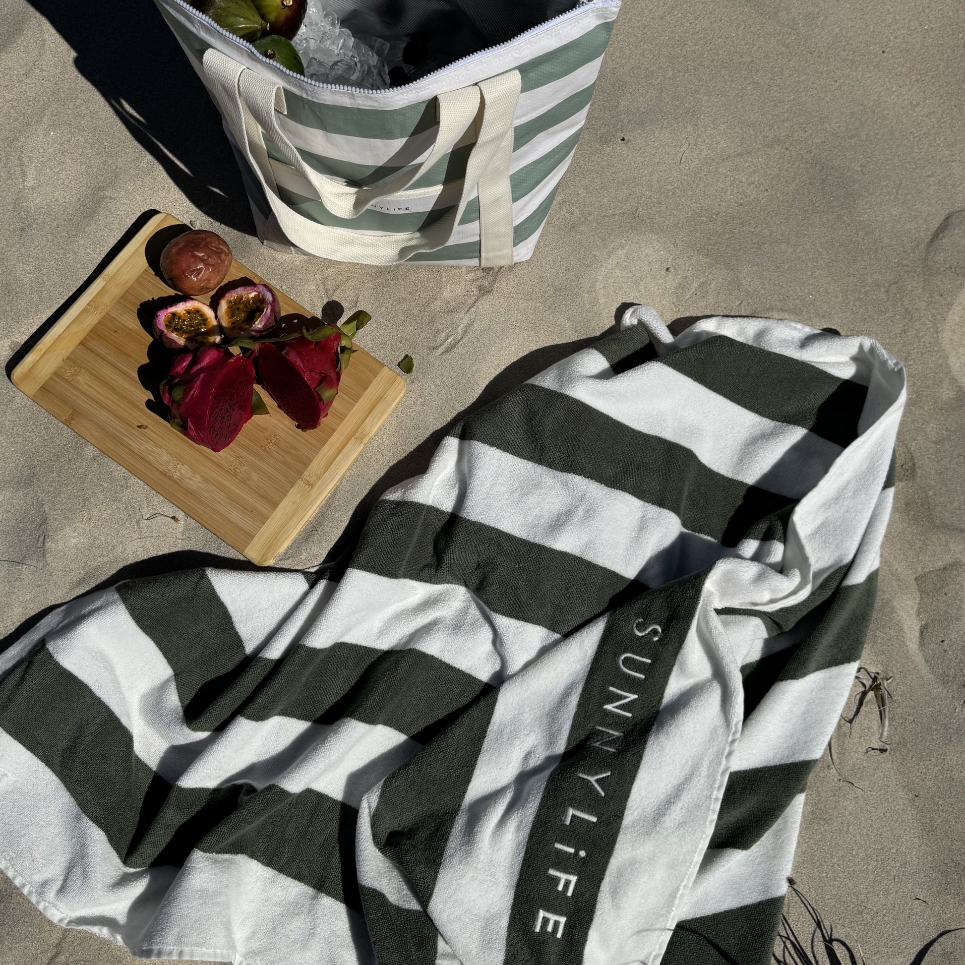 Torba chłodząca La Palma od Sunny Life w paski biało-zielone na plaży z ręcznikiem i owocami