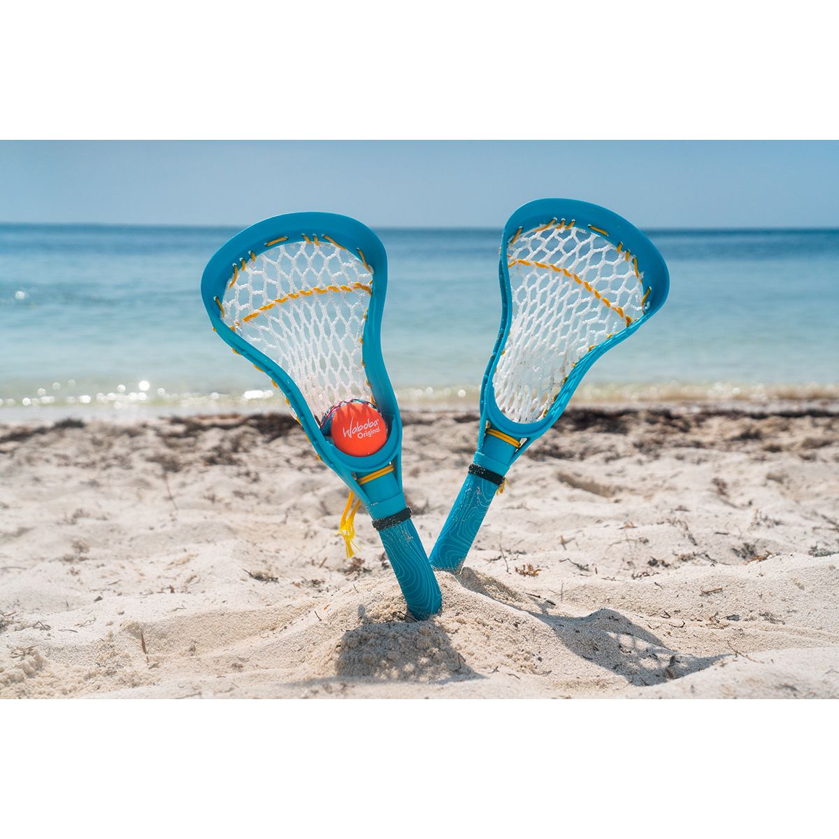 Zestaw do lacrosse dla dzieci marki Yamann na plaży z dwoma niebieskimi kijami i pomarańczową piłką