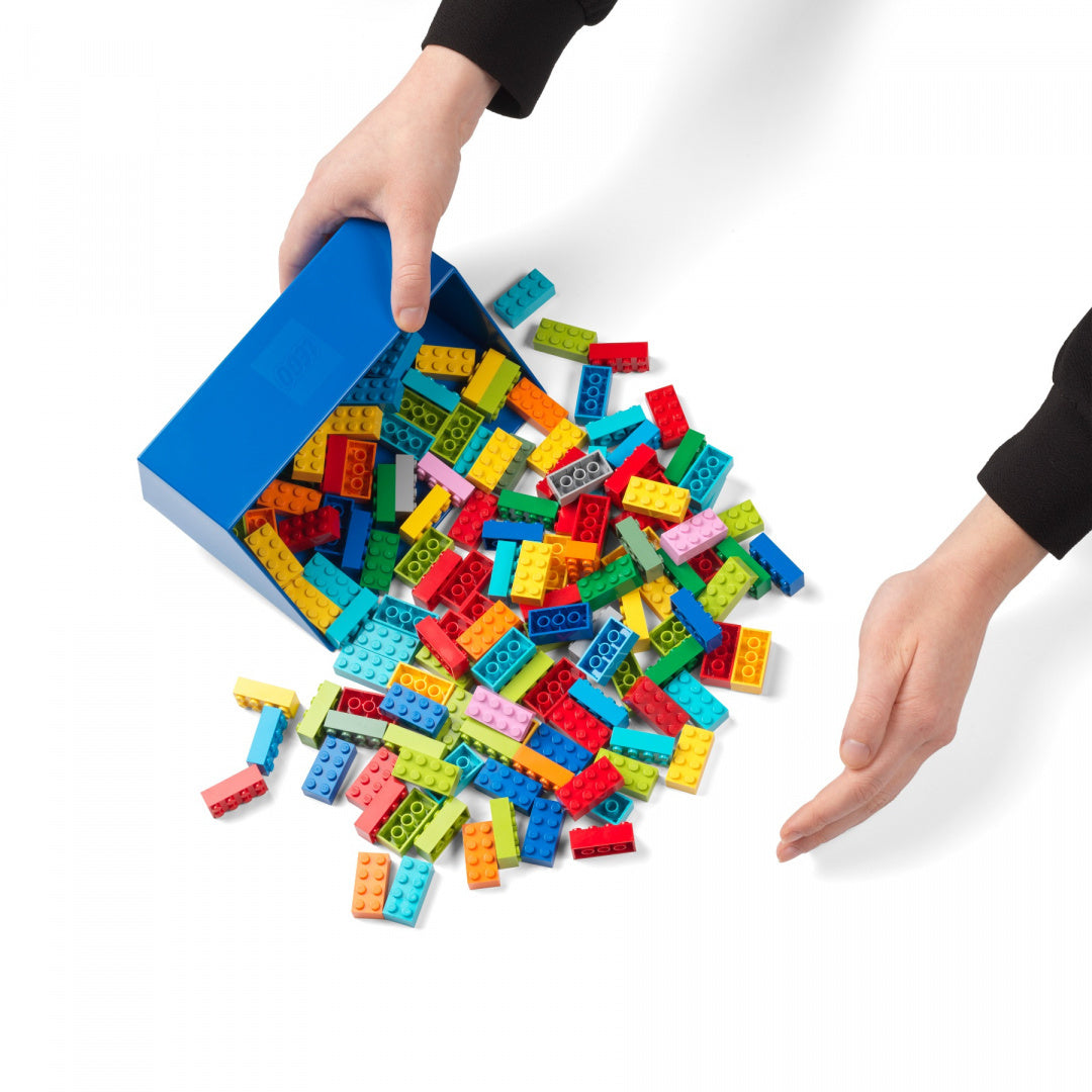 Ręce wysypujące zestaw szufelek LEGO z niebieskiego pojemnika na białym tle